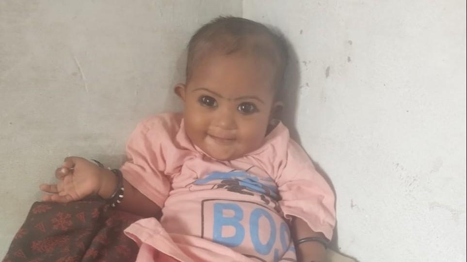 Hannah smiles at the camera while leaning in the corner of a room.  She is wearing a pink outfit with blue on the front.