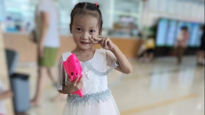 Zaria smiles at the camera and holds a peace sign next to her face. In the other hand, she holds a pink toy. She wears a beautiful sequined white dress.