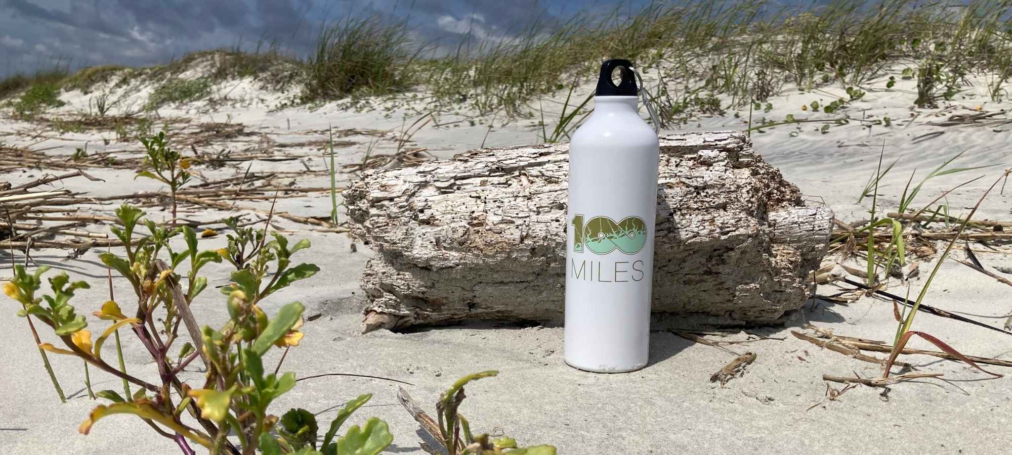 OHM branded white water bottle on white sand beach with driftwood behind