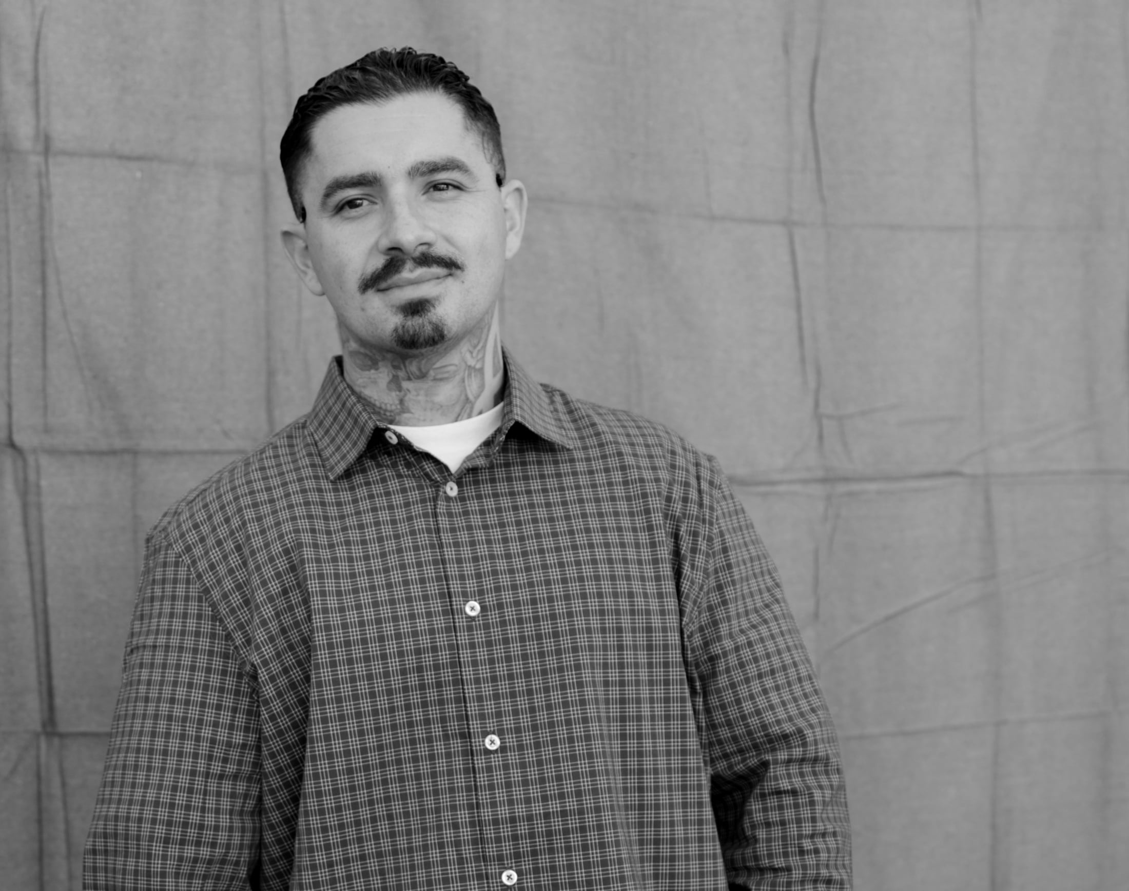 Latino man with tightly cropped hair, goatee and neck tattoos wearing a button-down shirt looks at the camera.