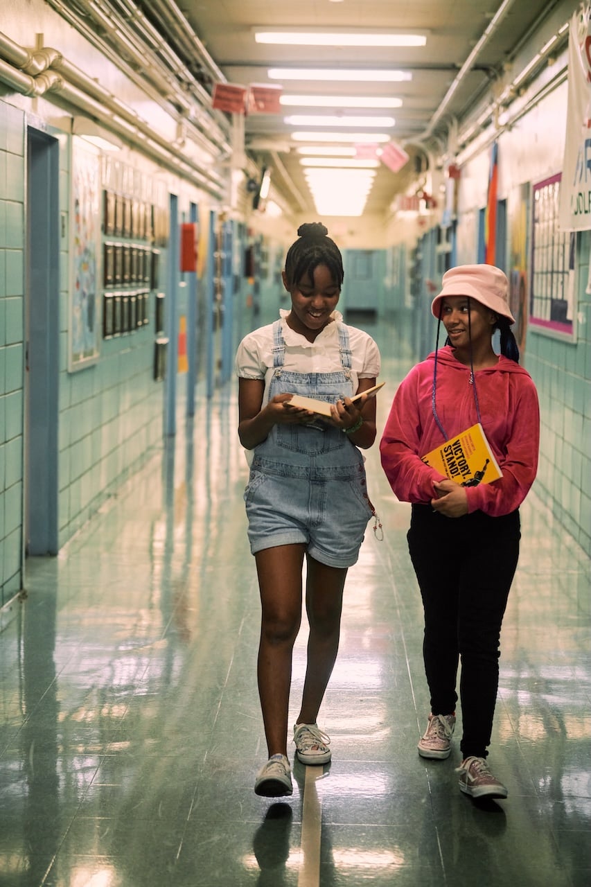 Two young girls walking down their school hallway while reading a book.